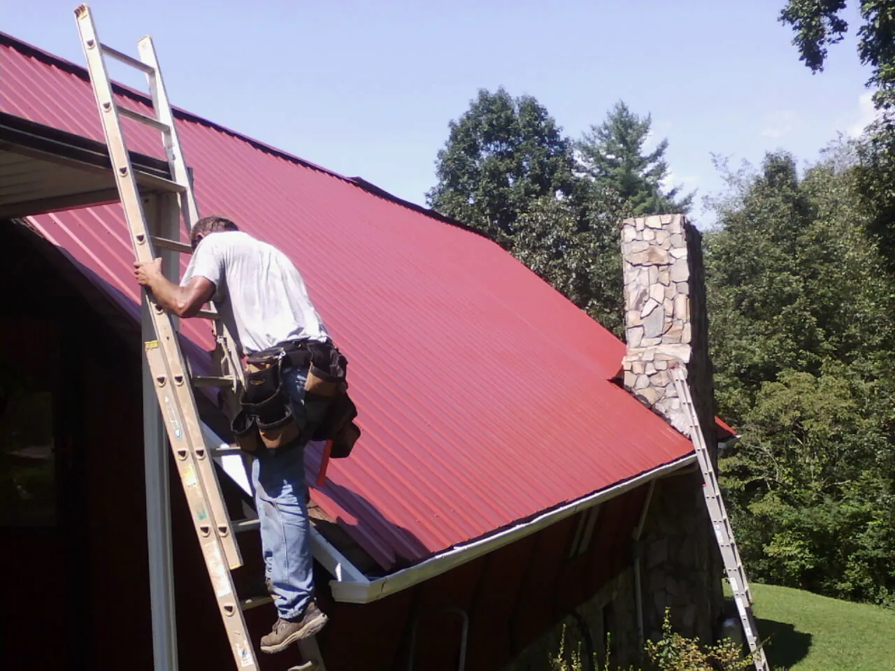 Roofer on a ladder beside a red metal roof and stone chimney — Soffit & Fascia Repair by Roofing Brothers in Lockhart